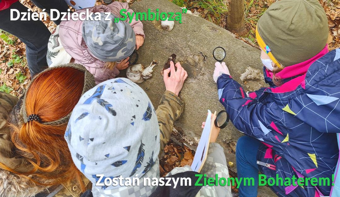 Baner przedstawiający akcję - Zostań Zielonym Bohaterem Symbiozy - w tle dzieci z edukatorem badający martwe drewno lupką